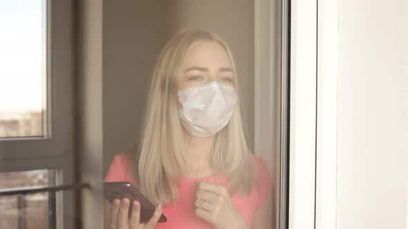 Girl in a Covid-19 Protection Mask Stands at Window and Is Sad.