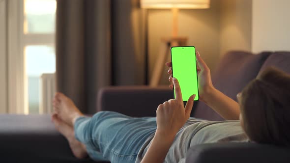 Woman at Home Lying on a Sofa and Using Smartphone with Green Mock-up Screen in Vertical Mode. Girl