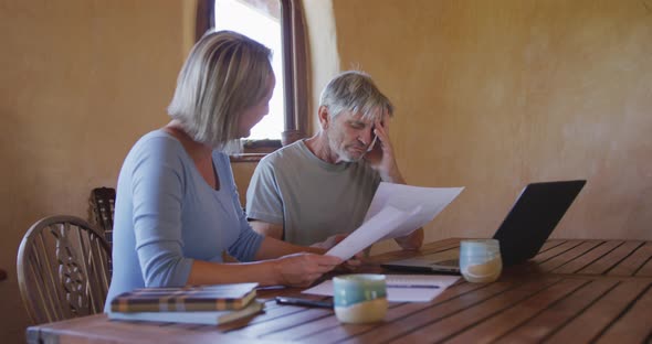 Senior caucasian couple using laptop and holding documents at table