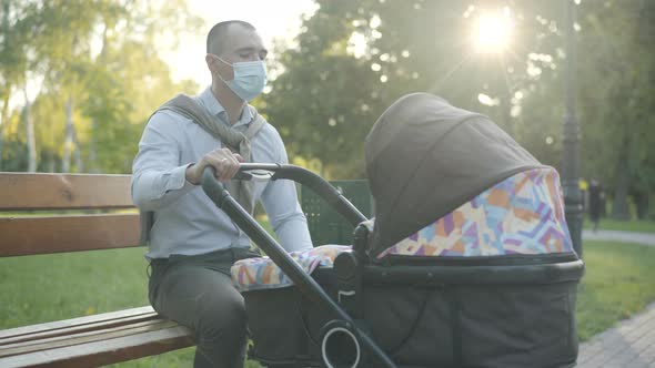 Young Confident Man in Covid-19 Face Mask Sitting in Sunlight and Rocking Baby Carriage in Summer