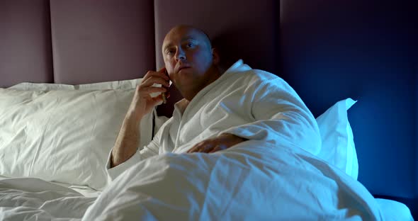 Adult Bald Man Who Recently Woke Up, Talking on a Mobile Phone, Lying on the Bed, Holding a