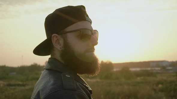 Bearded Brutal Trendy Guy in Black Glasses, Cap, Leather Jacket Is at Sunset. Fashionable Stylish