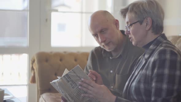 Old Couple, Man and Woman, Sitting on the Sofa with a Book and Talking and Smiling To Each Other