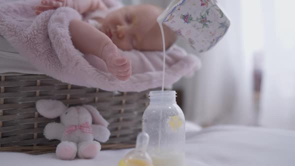 Pouring Milk in Baby Bottle with Blurred Infant Sleeping at Background in Baby Basket