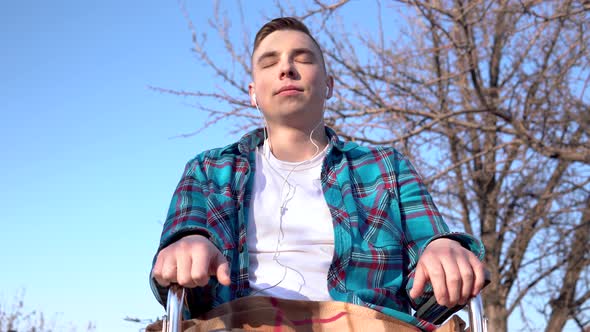 Young Man on a Wheelchair with Headphones. Disabled Man in Nature with Headphones in the Ears.