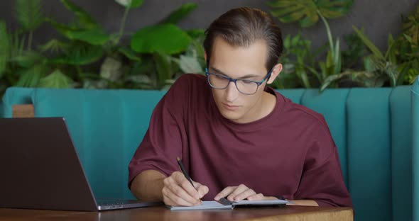 Handsome Young Man in Eyeglasses is Sitting at a Table in Cafe with a Laptop and Writes in Notepad