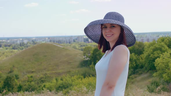 Young female in white dress and elegant hat on sunny day in hilly terrain.