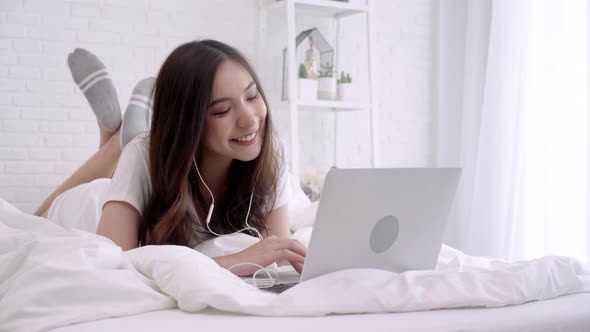 Asian woman playing computer while lying on the bed listening music with headphones .