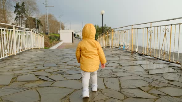 Handheld Camera Follows a Child a Girl in a Yellow Jacket or Coat Walking Forward Over a Cobbled