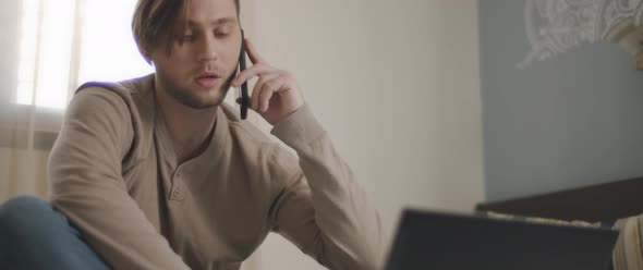 A man talking to someone on the phone while working with a laptop from home.