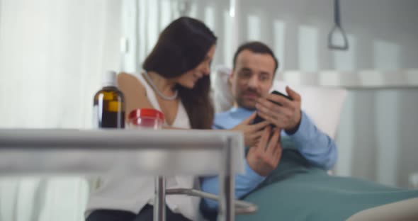 Young Woman Visiting Sick Husband in Hospital Watching Photo on Smartphone Together