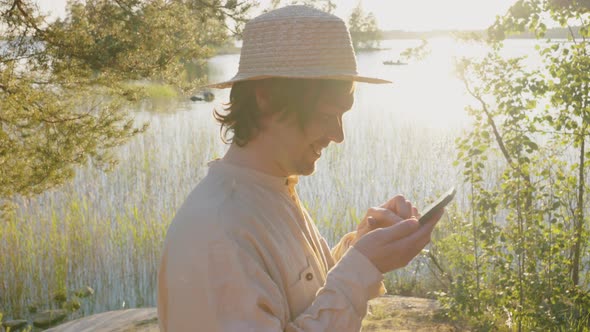 Man in Hat Tries to Get Cell Signal on Phone on River Bank