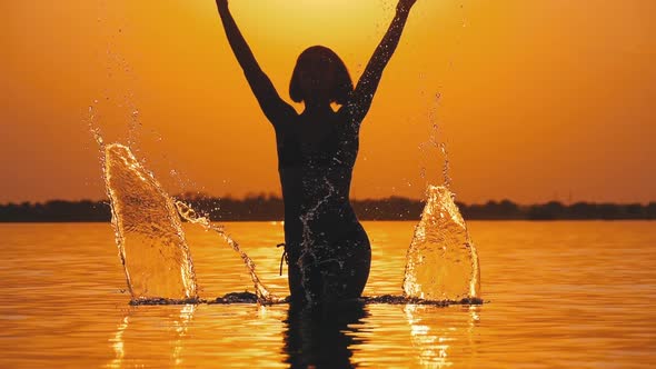 Silhouette of Woman at Sunset Raises Hands Up and Creating Splashes of Water. Slow Motion