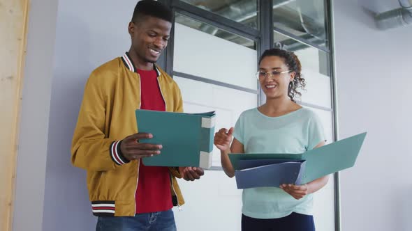 Happy diverse male and female creative colleague looking at documents in folders and talking