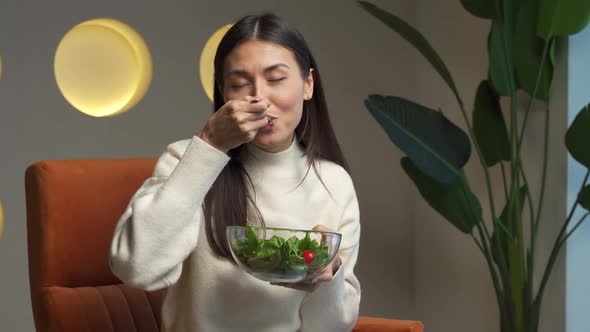 Cheerful Young Asian Woman Who Eats Fresh Salad and Smiles Sitting on a Chair in the Living Room