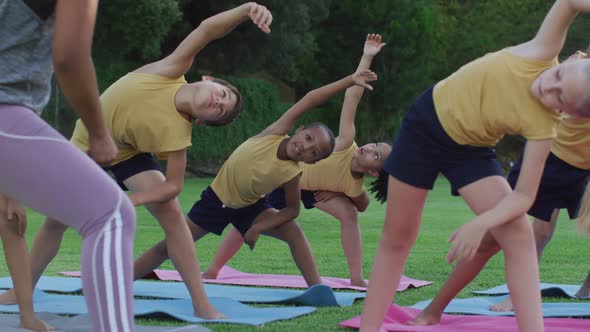 Mixed race female teacher showing diverse group of schoolchildren yoga stretching exercises outdoors