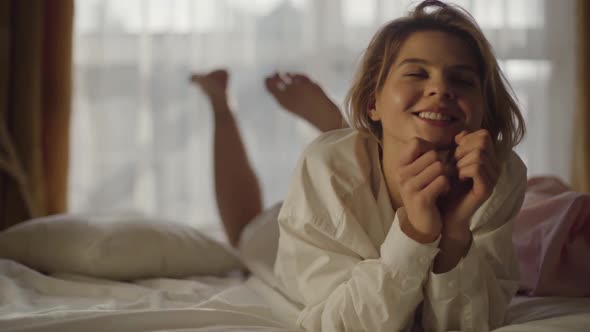 Portrait of Young Caucasian Woman Lying on Soft Bed and Shaking Legs. Smiling Girl in White Shirt