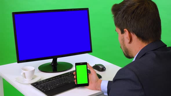 A Businessman Sits at a Desk with a Blue Computer Screen and Looks at a Smartphone - Green Screen