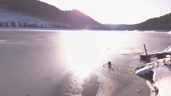 Descending aerial view of man neck deep in frozen mountain lake with sunlight shining on icy surface