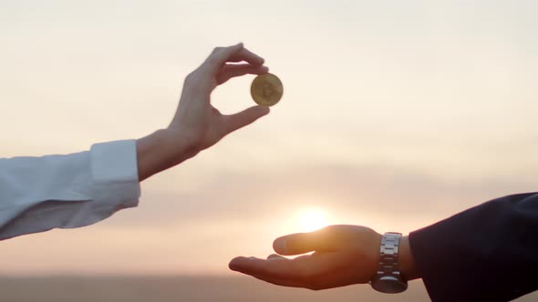 Closeup of Womans Hand Exchanging Bitcoin Gold Coin with Businessman Against Background of Sunset