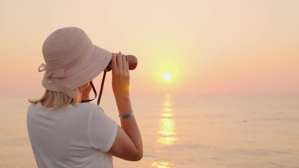 A Young Traveler in a Hat Looks Through Binoculars To the Pink Sea, Where the Sun Rises. Romance and