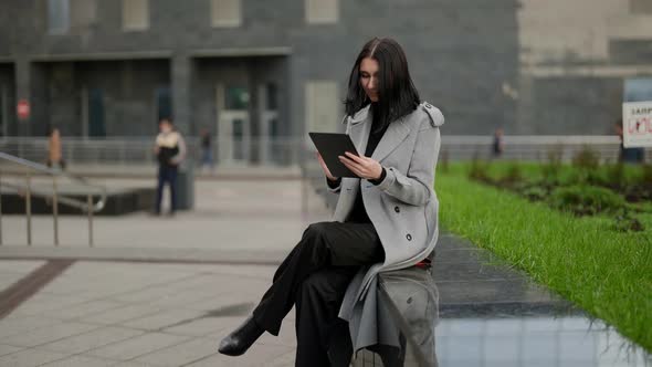 a Brunette in a Gray Coat Has Crossed Legs and is Sitting with a Tablet in Hands on a Stone Bench