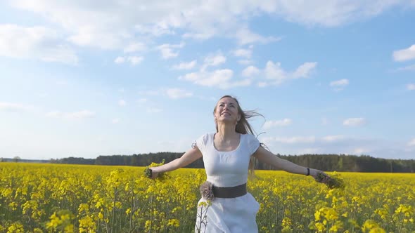 A Happy Girl in a White Dress Runs Among a Rapeseed Field on a Warm Spring Day. Slow Motion Mode. alt