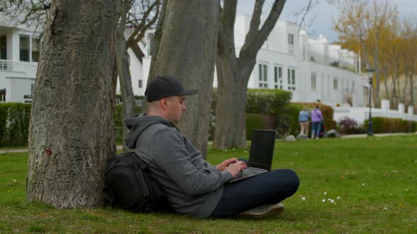 Young Man is Sitting in a Park in a Tourist Place Working on a Laptop