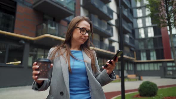 Formally Dressed Woman Walks Down the Street in a Business District with Coffee in Hand and Uses a