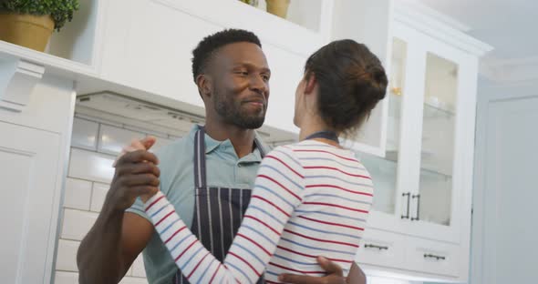 Happy diverse couple wearing blue aprons and dancing in kitchen