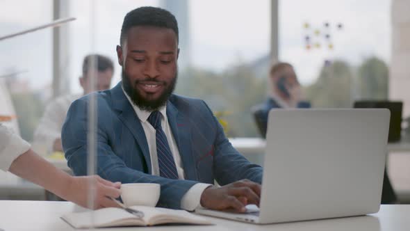 Secretary Bringing Coffee To Young African Businessman Working on Laptop