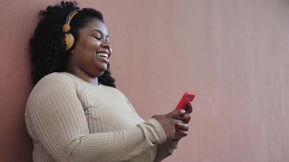 Happy Afro woman messaging online with mobile smartphone while listening music with headphones
