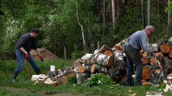 Elderly person and young man are chopping birch logs with an axes outdoors