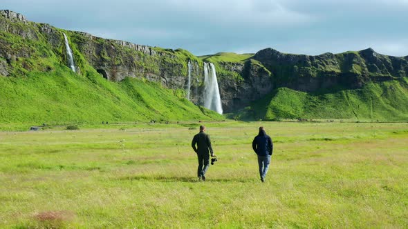 Two Tourists walking towards Seljalandsfoss and Gljufrabui waterfalls over a beautiful field of gras