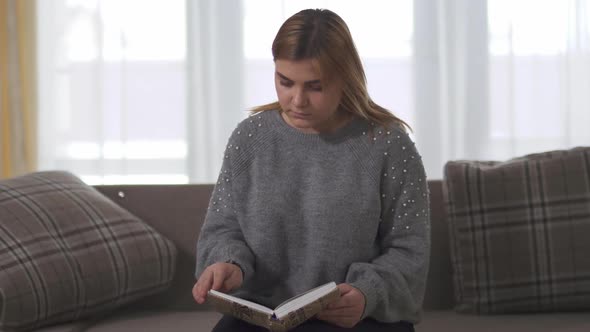 Plump Girl Reading Book Sitting at Sofa in Home