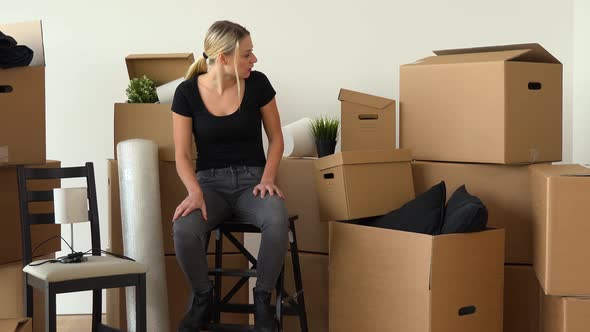 A Moving Woman Sits on a Chair in an Empty Apartment and Checks Cardboard Boxes Surrounding Her