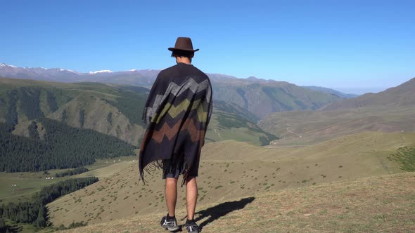 A Guy in a Hat and Poncho Stands on Edge of a Hill