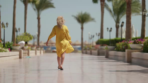 a Curly Blonde in Sunglasses and a Yellow Fluttering Dress Walks Away Against the Background of