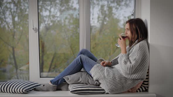 Young Woman Sitting at Windowsill with Glass of Red Wine