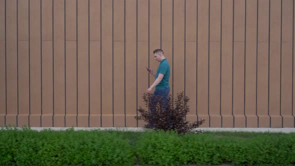 A Young Man Goes with a Phone in His Hand. A Man Walks Near the Building and Carries on