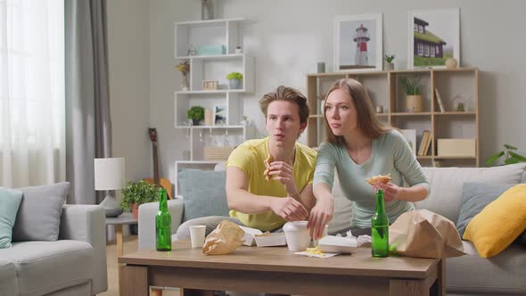 Young Couple Eating a Hamburger and Watching TV in the Living Room