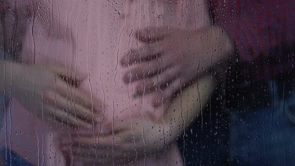 Pregnant Lady and Man Stroking Belly Behind Rainy Window, Waiting for Newborn