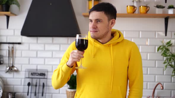 Relaxed Young Man Poses with Glass of Red Wine Standing at Kitchen Table