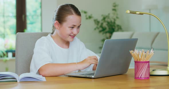Cute Teen Girl Sits at the Table with Laptop and Writes Smiling Notes and Pencils and a Lamp are on alt