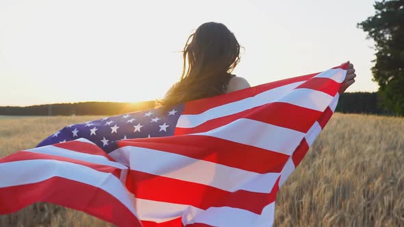 American girl running with open arms over wheat field and holding USA flag