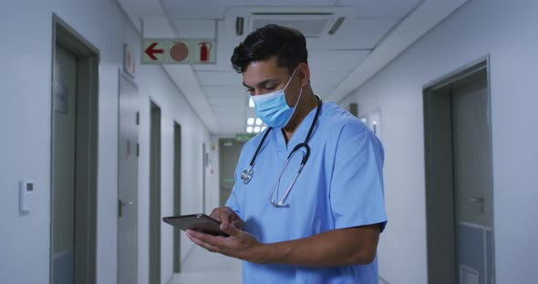 Mixed race male doctor wearing face mask standing in hospital corridor using tablet