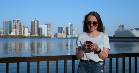 Beautiful Girl with Long Hair in Sunglasses Using Smartphone App at Sunset River Quay Near