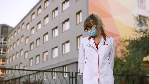 Exhausted and Depressed Female Doctor or Nurse in Surgical Face Mask Sits at Hospital Entrance