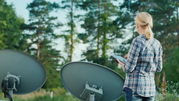 Rear View of Woman Tunes Satellite Dishes Outdoors, Uses Tablet