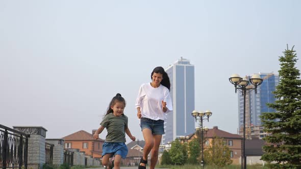 Smiling Asian Girl with Mother Runs Along City Embankment